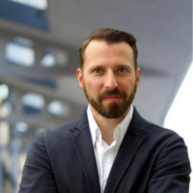 Erik Bade M. Sc. Mann mit Bart und Anzug, lächelt vor modernen Architektur im Hintergrund.